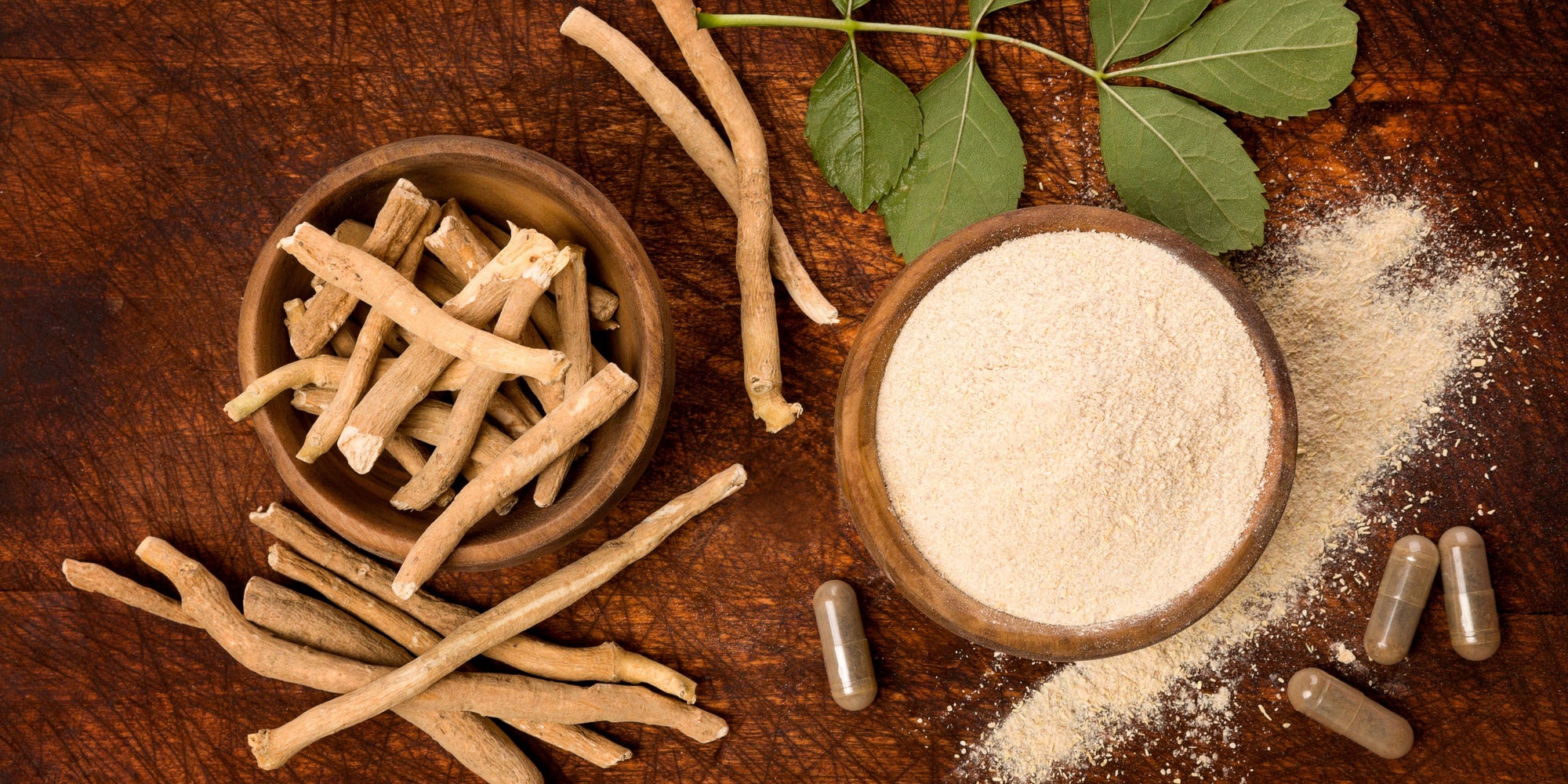 
                  wooden bowl of ashwagandha roots next to a wooden bowl of powder, capsules and leaves
                