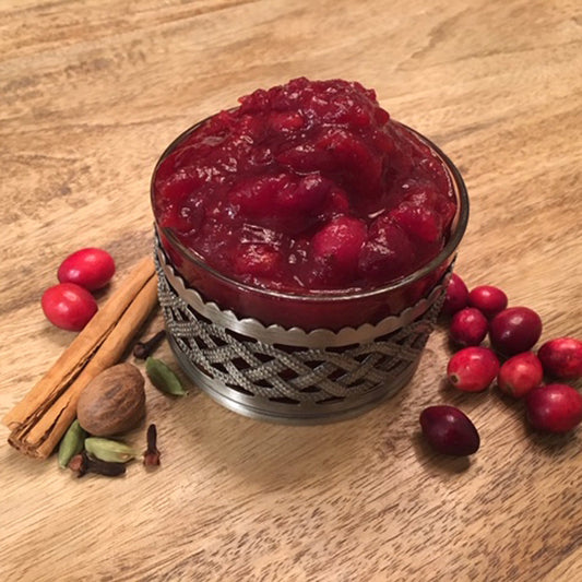 A small glass of spiced cranberry sauce next to cloves, nutmeg, a cinnamon stick and cranberries 