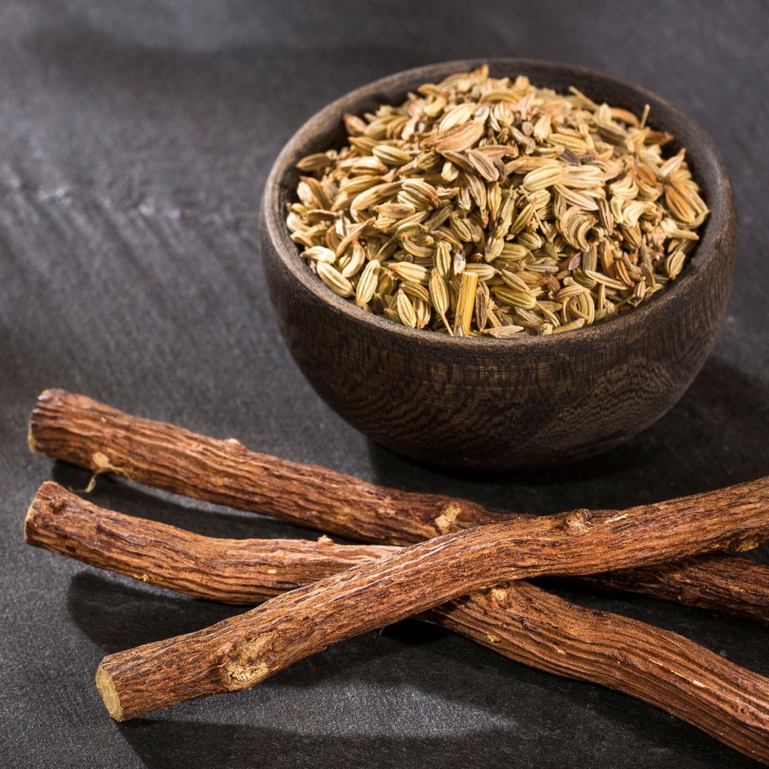 Small bowl of fennel seeds next to 3 sticks of licorice roots against dark background