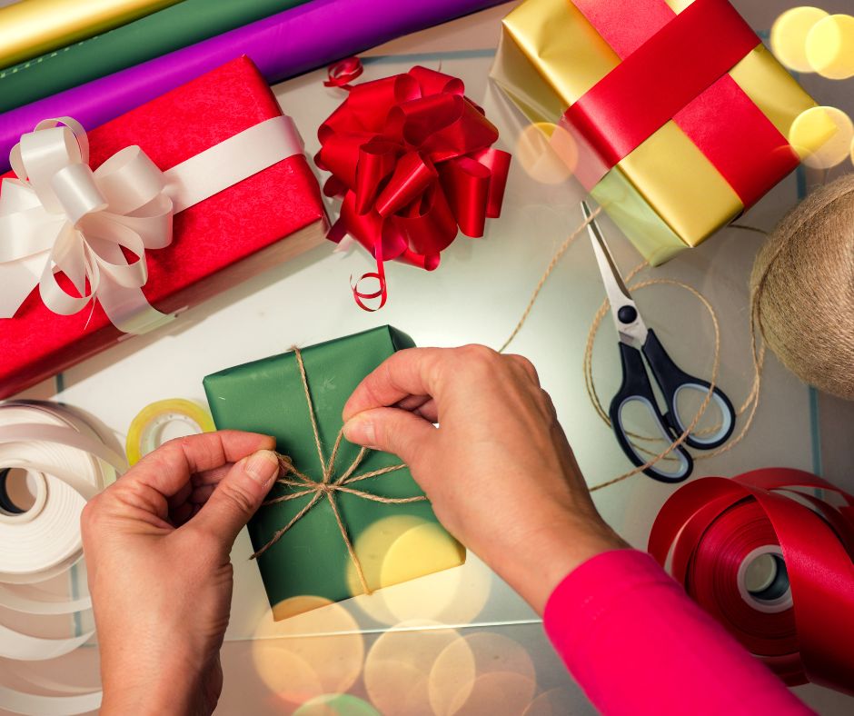 A pair of hands wrapping a green gift with twine surrounded by wrapping paper, wrapped gifts, tape, and scissors. 