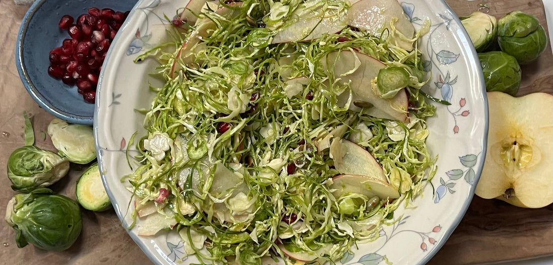 Brussel sprout salad in a bowl, surrounded by brussel spouts, pomagranate seeds and apple slices  