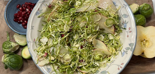 Brussel sprout salad in a bowl, surrounded by brussel spouts, pomagranate seeds and apple slices  