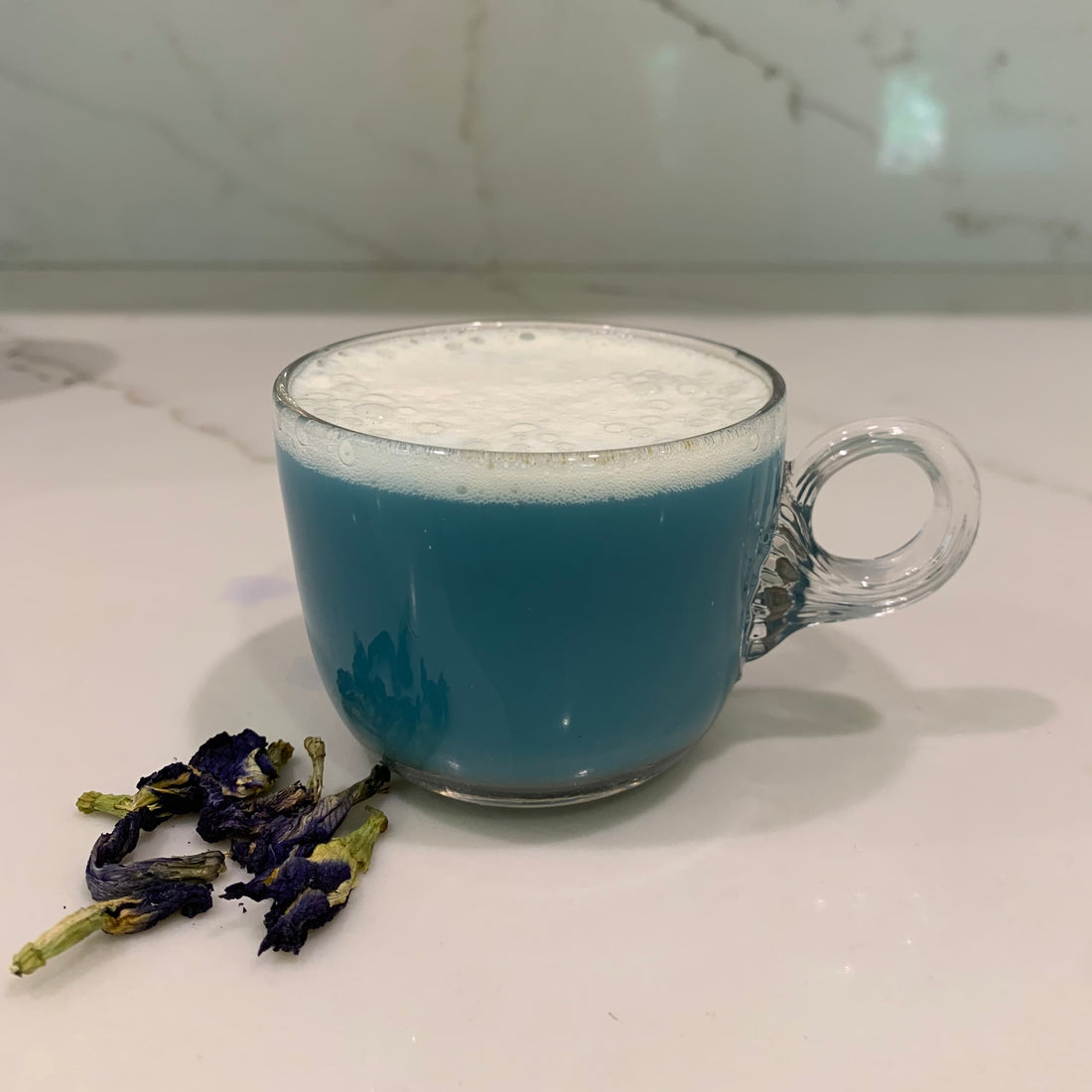 a clear mug with blue moon tea on a white counter with a couple of Pea flowers next to the cup.