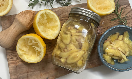 Ginger strips in a jar, on a cutting board with lemons, herbs, a bowl of ginger, and a wooden juicer