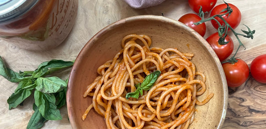 Jar of sauce, next to pasta in sauce and tomatoes on a wooden cutting board