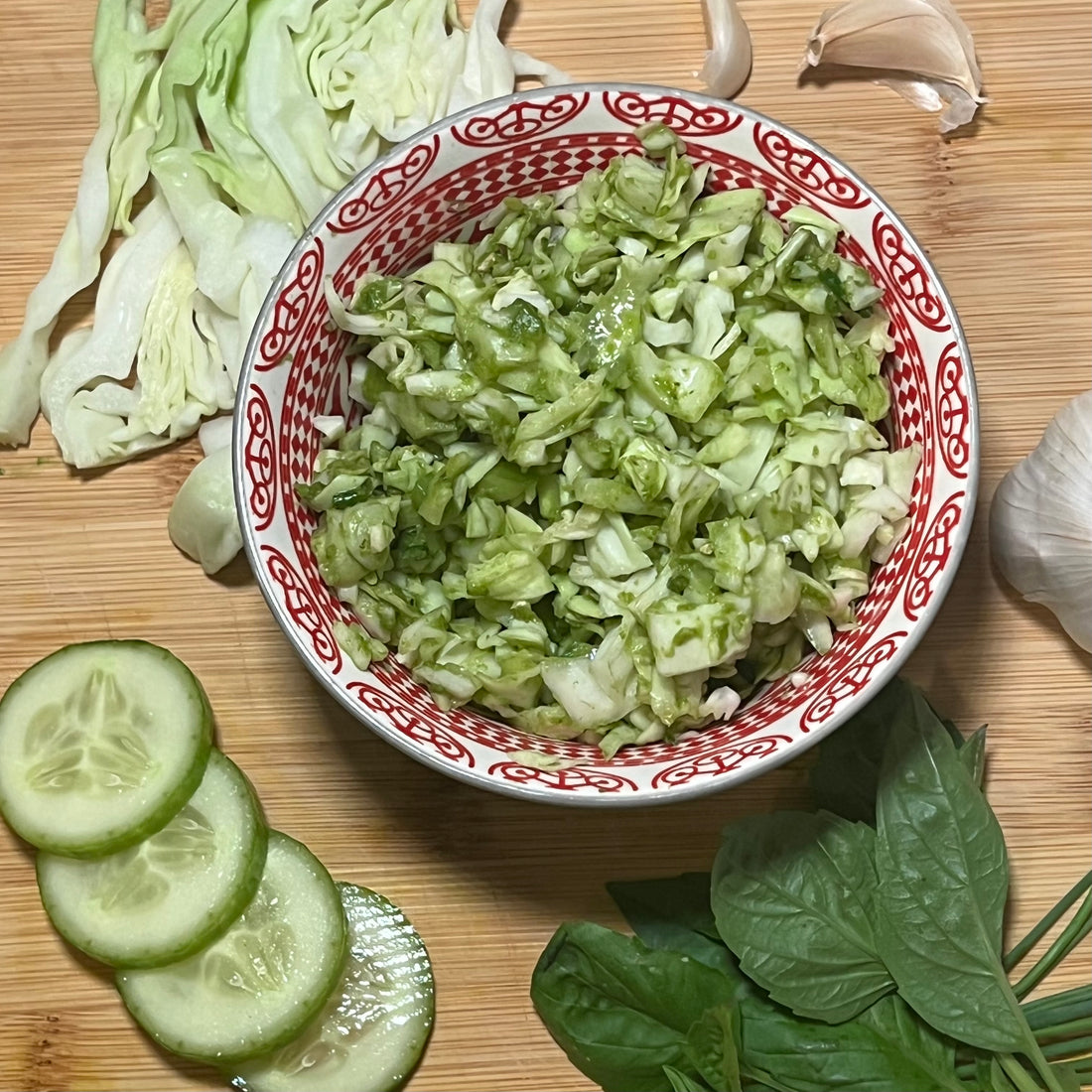 Completed salad in bowl surrounded by ingredients, cabbage, basil leaves, chives, cucumber and garlic 