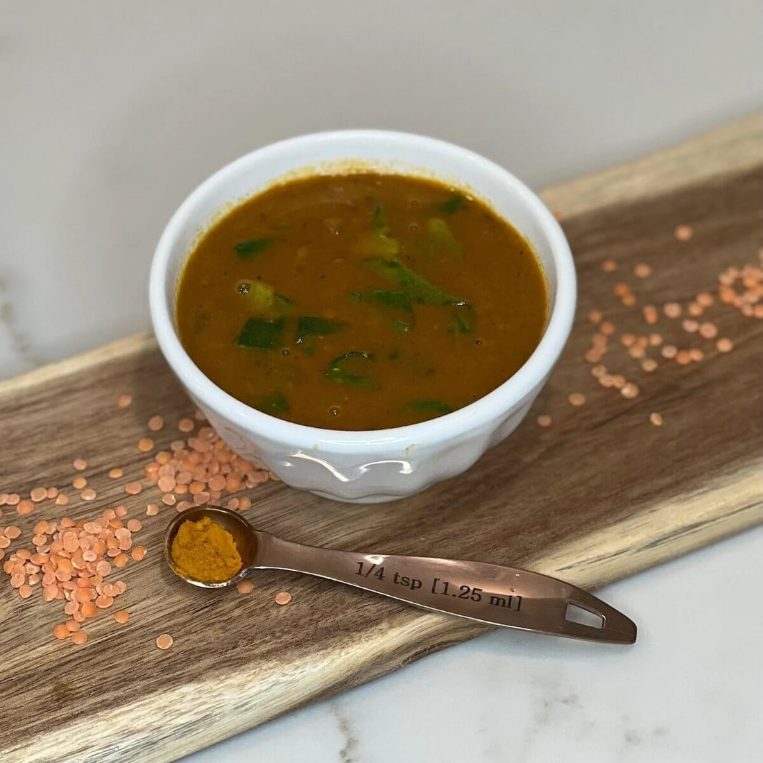 small bowl of lentil soup on a cutting board with red lentils spread around and a teaspoon next to the bowl with turmeric powder in it