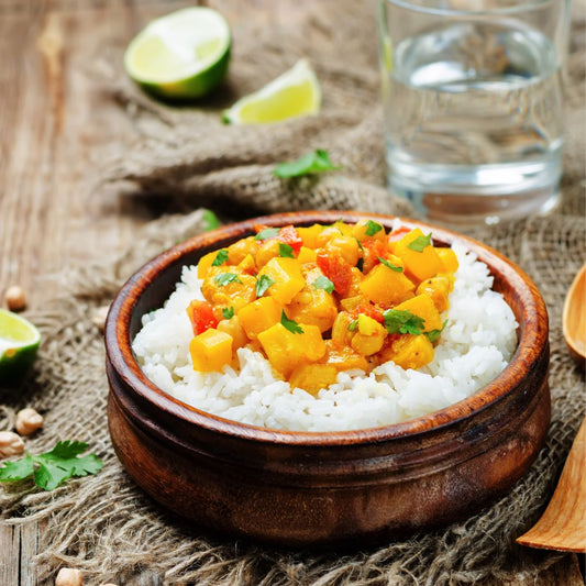A wooden bowl with rice and curry on table with a cup of water, wooden spoon, and sliced limes