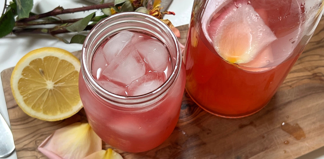 A close up of a glass of rose water lemonade next to pitcher and lemon sliced in half, on wooden board.