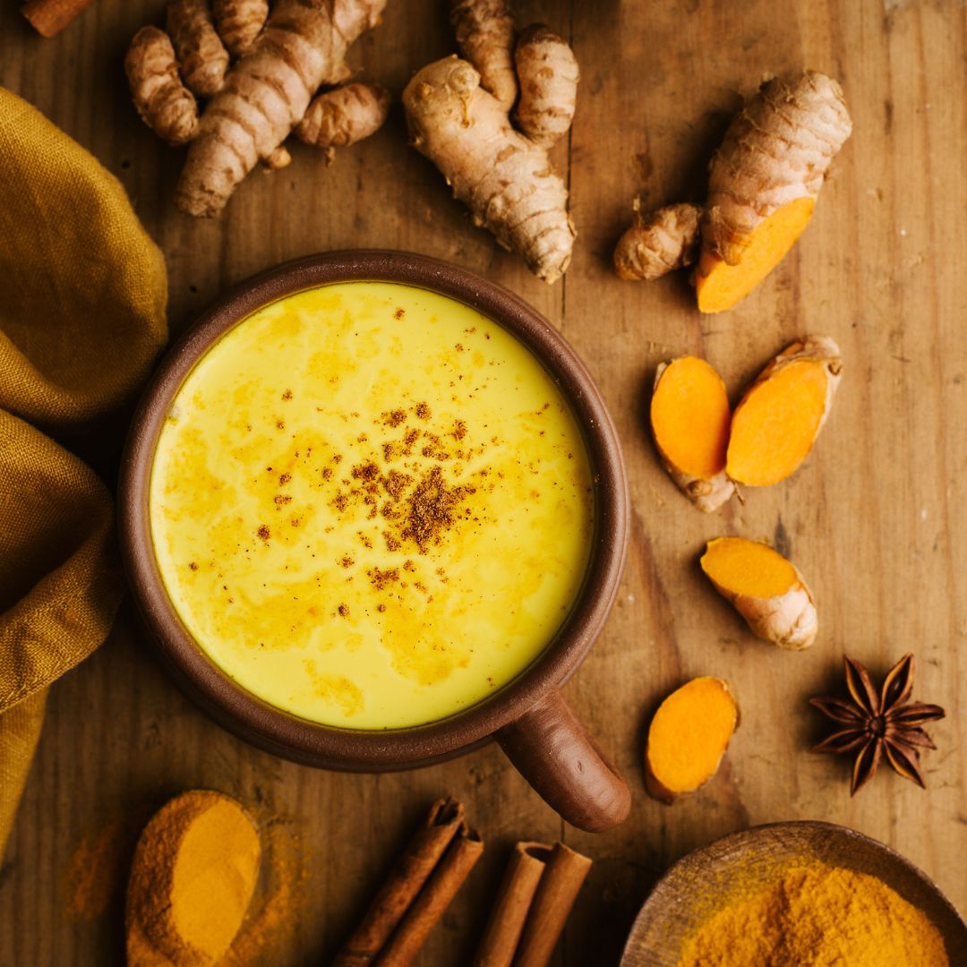 herbal roots turmeric bottle with a bottle of organic blue agave syrup and a cup of milk alternative sitting on a cutting board along with fresh cut ginger, black pepper seeds, turmeric and coconut oil