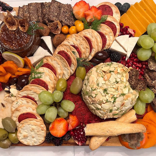 charcuterie board filled with crackers, green grapes, green and black olives, homemade vegan cheese ball, raw figs and homemade fig preserve, pomegranate seeds, sliced persimmon, flax seed crackers, raspberries, raisins, bread sticks, and sprigs of green