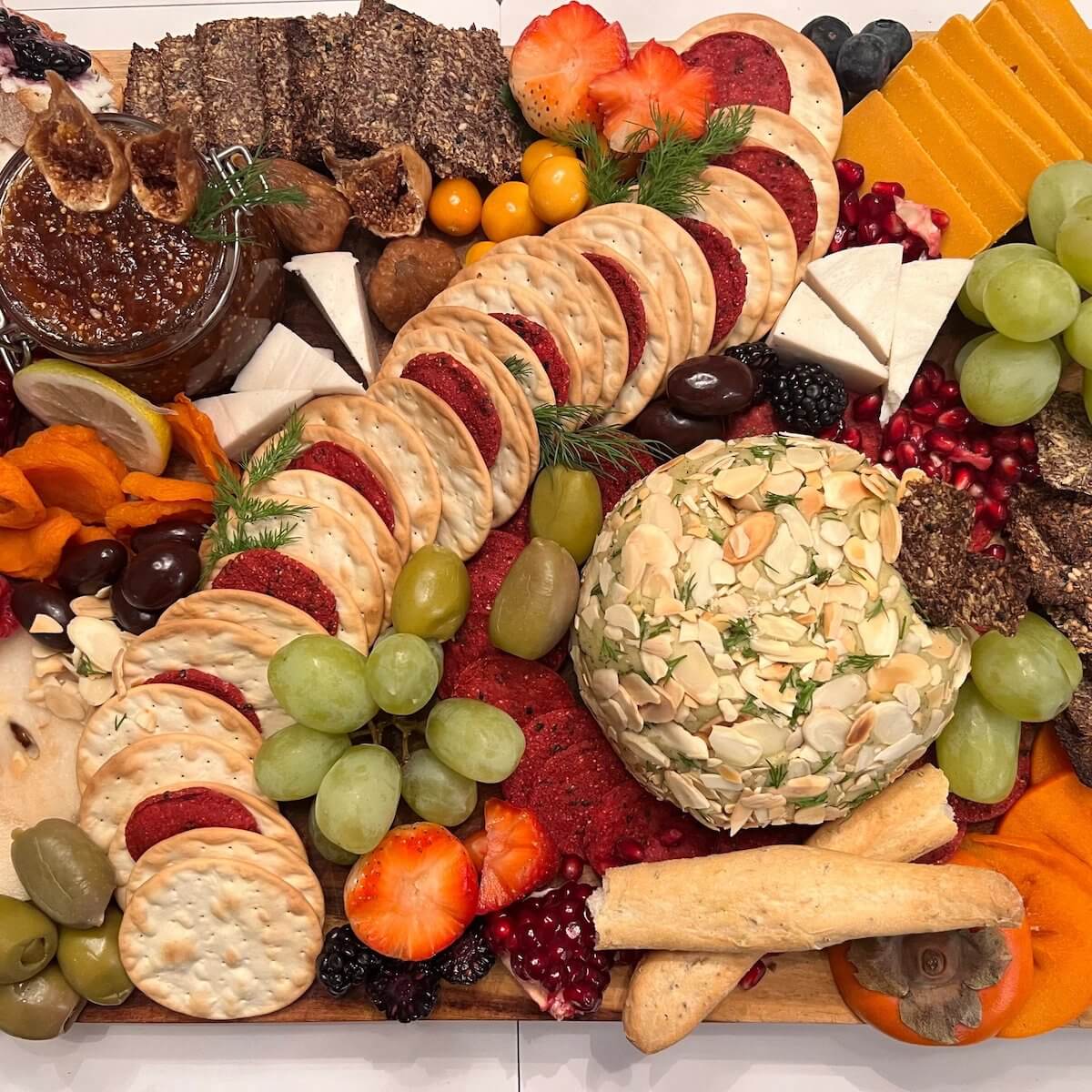 
          charcuterie board filled with crackers, green grapes, green and black olives, homemade vegan cheese ball, raw figs and homemade fig preserve, pomegranate seeds, sliced persimmon, flax seed crackers, raspberries, raisins, bread sticks, and sprigs of green
        