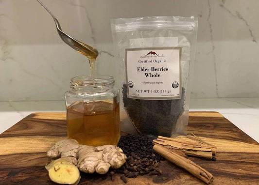 A glass jar with syrup next to a bag of whole elderberries, ginger root, clove, and cinnamon on a wooden board