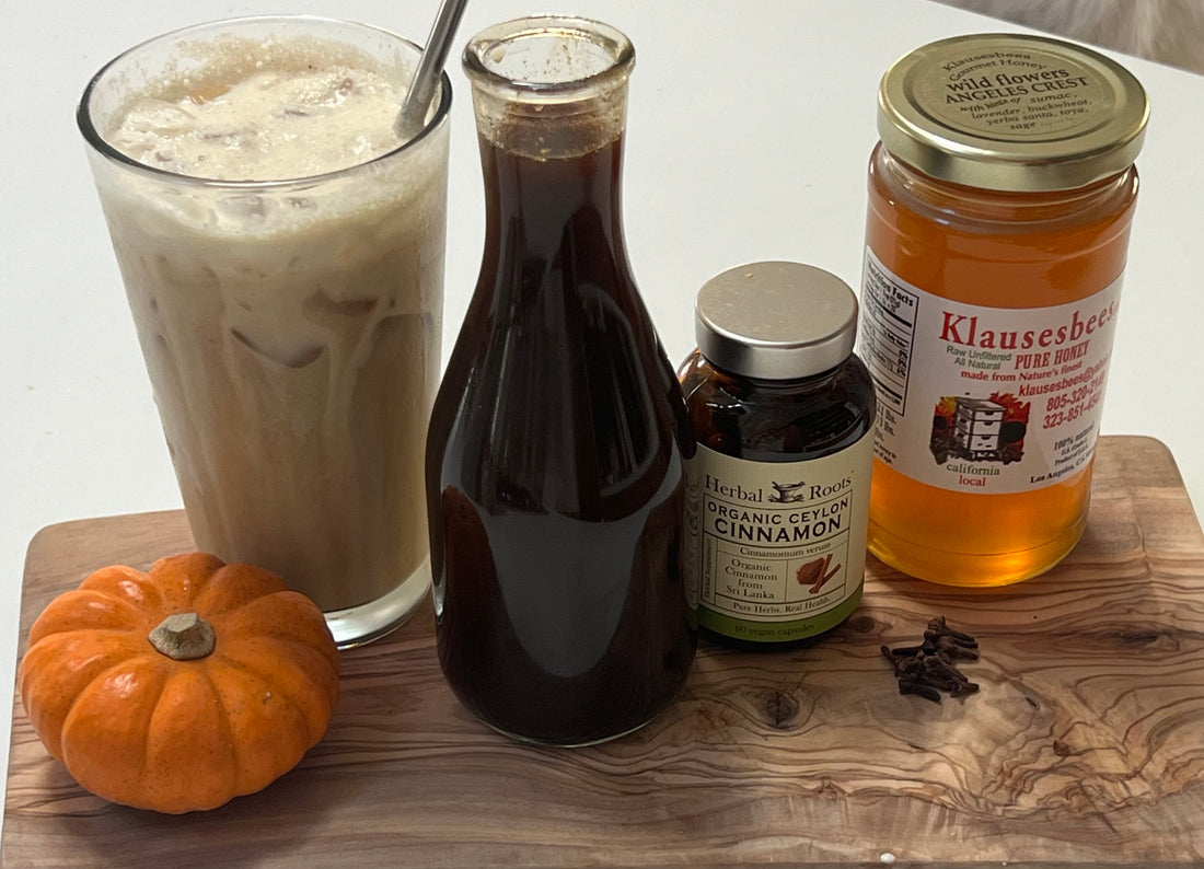 Cutting board with a full glass of iced coffee, a jar of homemade pumpkin spice syrup, a bottle of Herbal Roots Ceylon cinnamon capsules, jar of honey