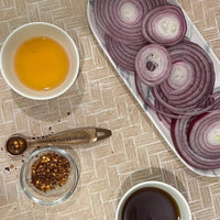 top view of a plate of sliced onions, small cup of vinegar, small cup of red pepper flakes, and a measuring spoon