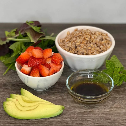 pile of spinch, bowl of cooked farro, bowl of cut strawberries, sliced avocado, sprig of mint