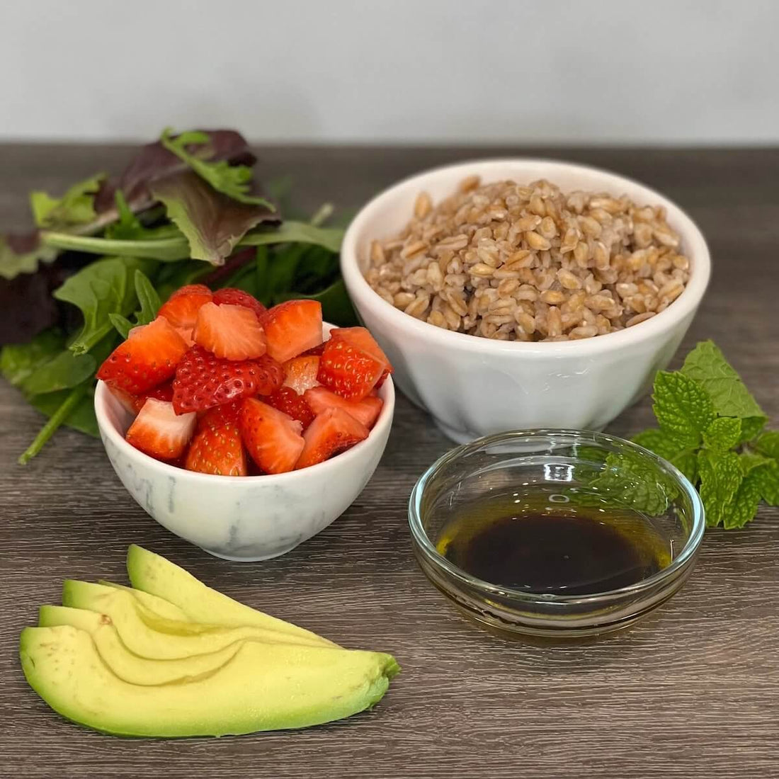 pile of spinch, bowl of cooked farro, bowl of cut strawberries, sliced avocado, sprig of mint