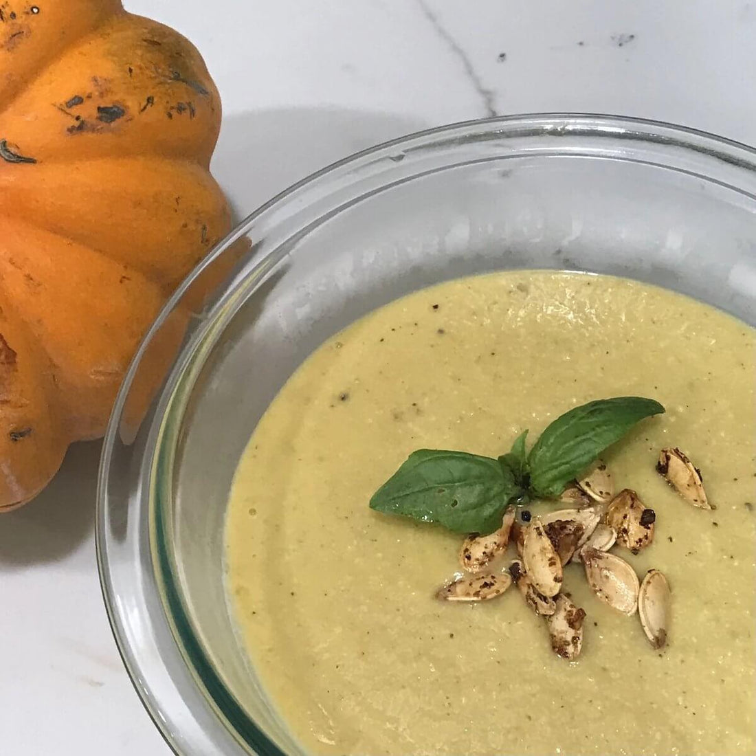 clear bowl of cream colored pumpkin soup with roasted pumpkin seeds and basil, next to a pumpkin