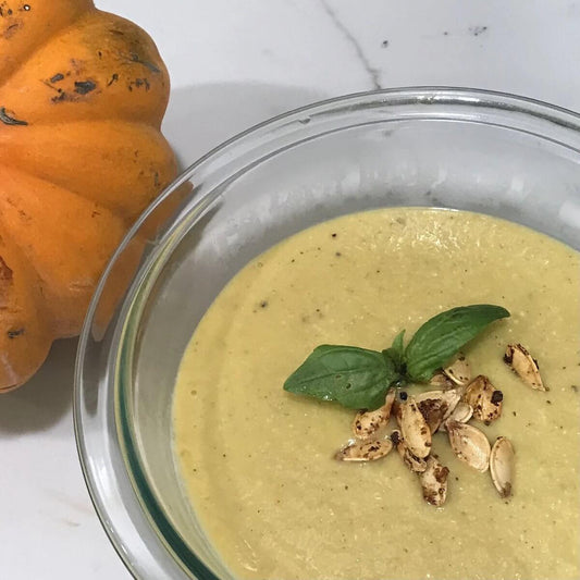 clear bowl of cream colored pumpkin soup with roasted pumpkin seeds and basil, next to a pumpkin